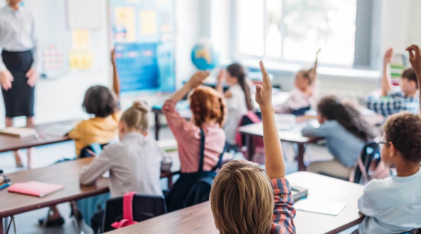 Dzieci siedzą w ławkach szkolych i zgłaszają się do odpowiedzi