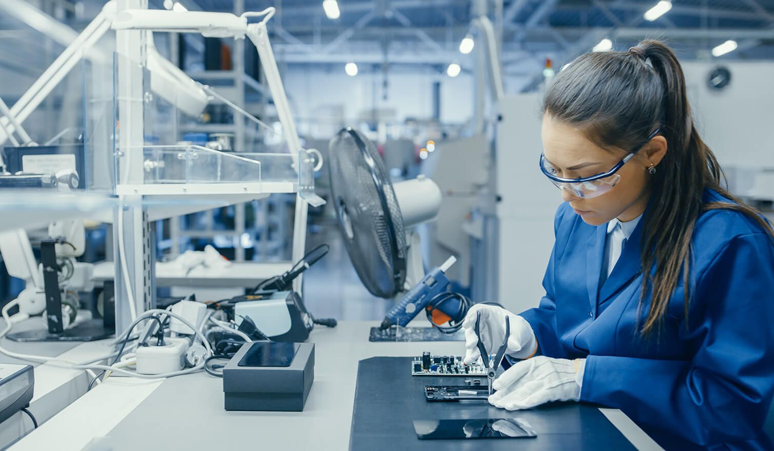 A woman in a blue uniform is carefully operating electronic equipment in a well-lit production environment
