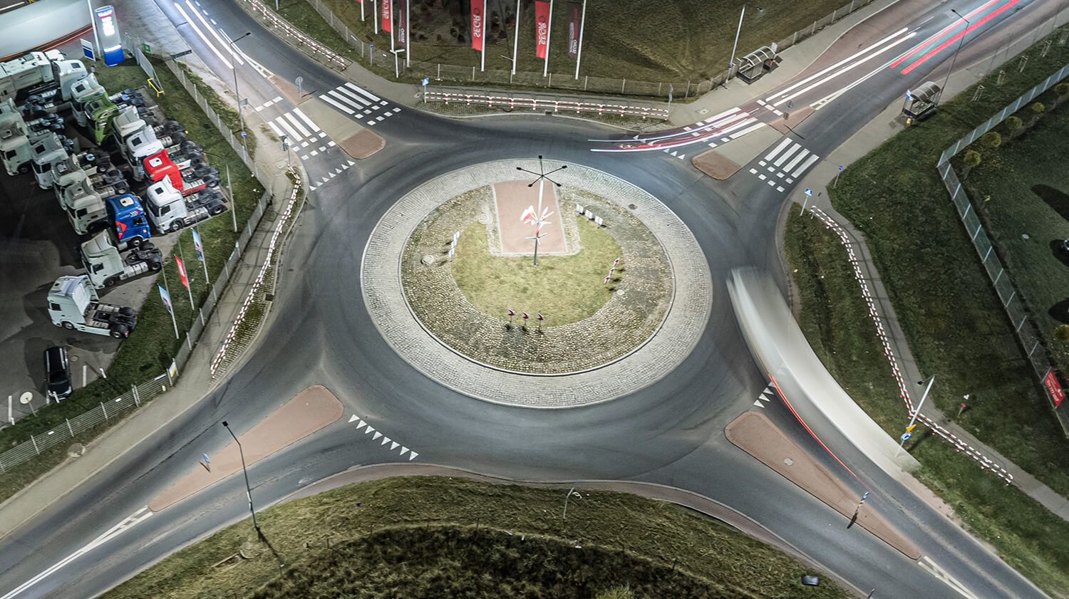 A night-time aerial view of a roundabout lit by street lamps, showing traffic and road markings indicating the direction of travel.